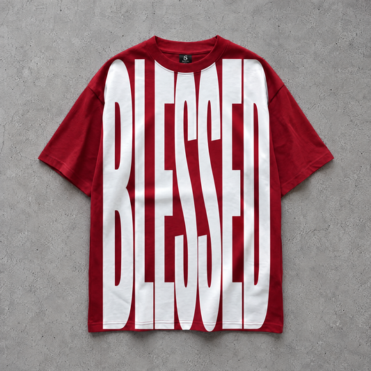A red oversized boxy short-sleeve t-shirt with the word 'BLESSED' printed in large white block letters across the front, laid flat on a textured gray concrete background.