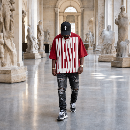 A man walking down a grand, classical hall lined with stone statues, wearing a red t-shirt with 'BLESSED' printed in white, a black baseball cap with a white cross, distressed black jeans, and sneakers.