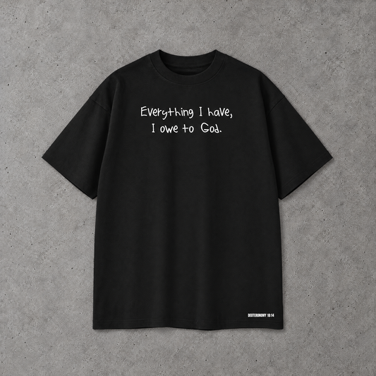 A black  short-sleeve t-shirt with the white handwritten text 'Everything I have, I owe to God.' printed across the chest, laid flat on a textured gray concrete background.