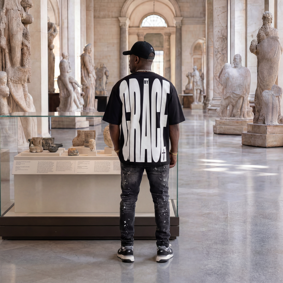 A man stands with his back to the camera in a grand museum hall, wearing a black t-shirt with 'BY GRACE' printed in large white letters on the back, paired with dark jeans and a black cap.