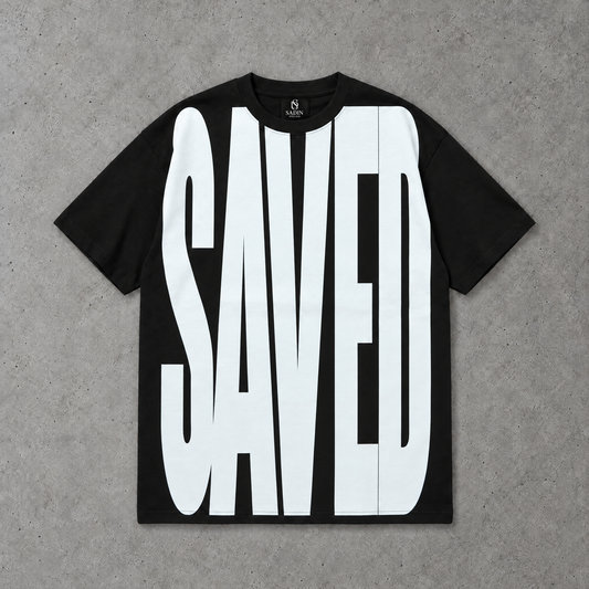 A black oversized box short-sleeve t-shirt with a large white graphic text print reading 'SAVED' across the front, laid flat on a textured gray concrete background.