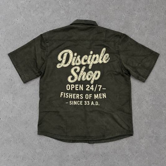An ovintage black short-sleeve button-up shirt shown from the back, laid flat on a textured gray concrete surface. The back features a large graphic print in cream and gold glittery lettering that reads 'Disciple Shop' with the phrases 'OPEN 24/7-', 'FISHERS OF MEN', and '- SINCE 33 A.